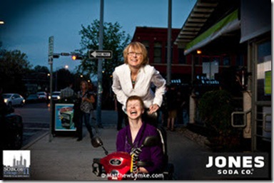 Liz Strauss standing on the back of Glenda Watson Hyatt's scooter